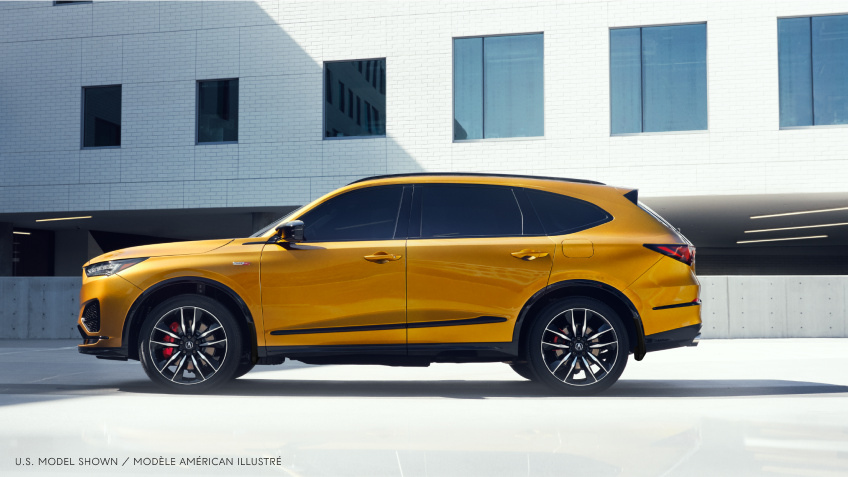 Plan étendu latéral d’un MDX Type S de couleur or à l’arrêt dans un stationnement situé à l’extérieur d’un bâtiment moderne de couleur blanche au style minimaliste.