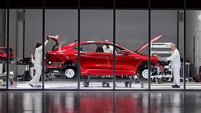 Passenger side view of a red Acura frame on a hoist in a glass-walled room. Acura engineers in white lab uniforms are seen inspecting the vehicle. Vue du côté passager d’un cadre Acura rouge sur un pont élévateur dans une pièce aux parois en verre. Ingénieurs Acura en sarraus blancs qui inspectent le véhicule.