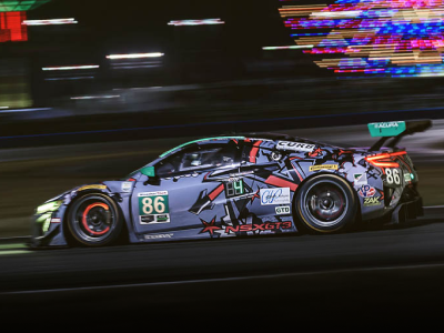 Driver side shot of a purple, sponsorship wrapped Acura NSX GT3 driving at high speed on a racetrack at nighttime. Vue du côté conducteur d’une Acura NSX GT3 recouverte de logos de commanditaires roulant à haute vitesse sur un circuit de course la nuit.