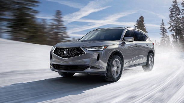 Front, side-angled view of a silver Acura MDX kicking-up snow on a wintery road with blue sky and trees blurred by the speed of the vehicle. // Vue avant latérale d’un Acura MDX de couleur argent roulant à pleine vitesse qui soulève la neige sur son passage avec à l’arrière-plan un ciel bleu et des arbres.