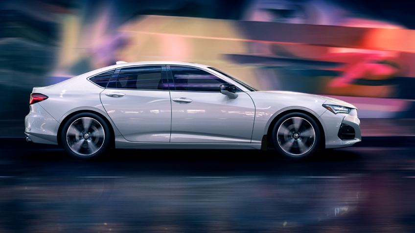 Passenger side view of a white Acura TLX driving at night - red, purple and yellow lights are blurred in the background by the motion of the car. // Vue du côté passager d’une Acura TLX blanche circulant la nuit avec en arrière-plan des lumières floues rouges, violettes et jaunes.