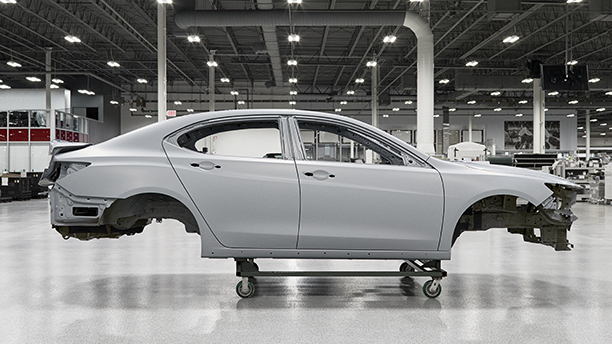 Passenger side view of a white vehicle frame hoisted on a dolly inside the Acura Performance Manufacturing Centre Vue du côté passager d’un cadre de véhicule blanc sur un chariot à l’intérieur du Performance Manufacturing Centre d’Acura.