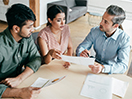 Three people sit around a table discussing paperwork. / Trois personnes sont assises autour d'une table pour discuter au sujet de documents.